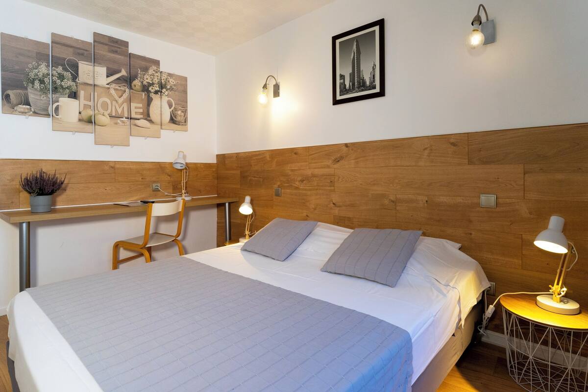 Un dormitorio con cabecera de madera, cama con ropa de cama gris, dos lámparas de noche, un escritorio con una silla y una obra de arte en la pared con la palabra 