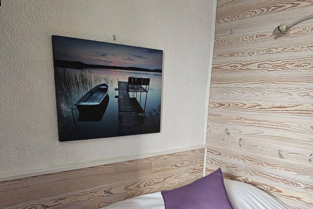 Un dormitorio con cama, almohada morada, control remoto y un cuadro de un muelle y un bote.