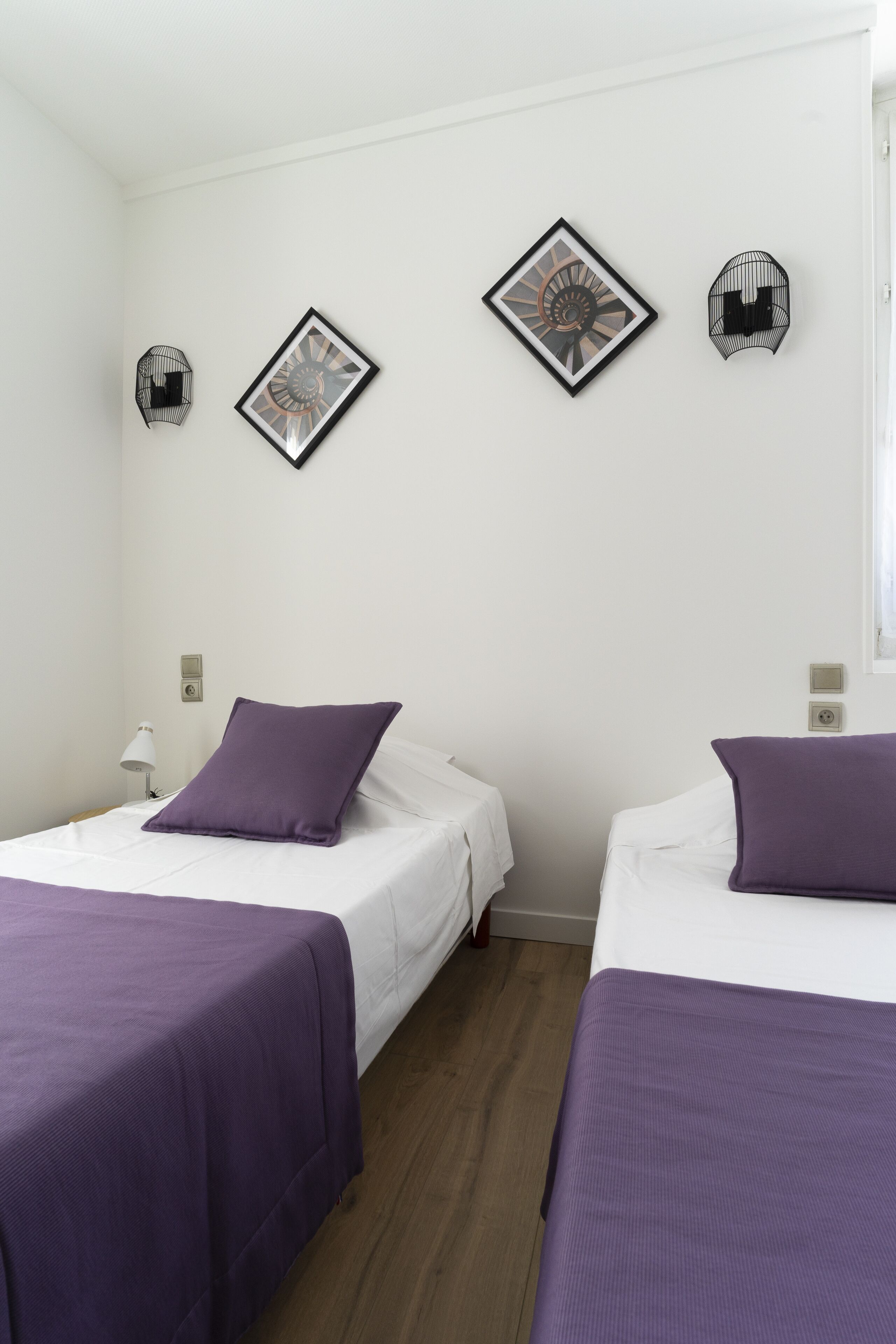 Habitación de hotel con dos camas, cada una con cubrecama morado, ropa de cama blanca y una mesita de noche pequeña con lámpara. Hay obras de arte colgadas en la pared sobre las camas.