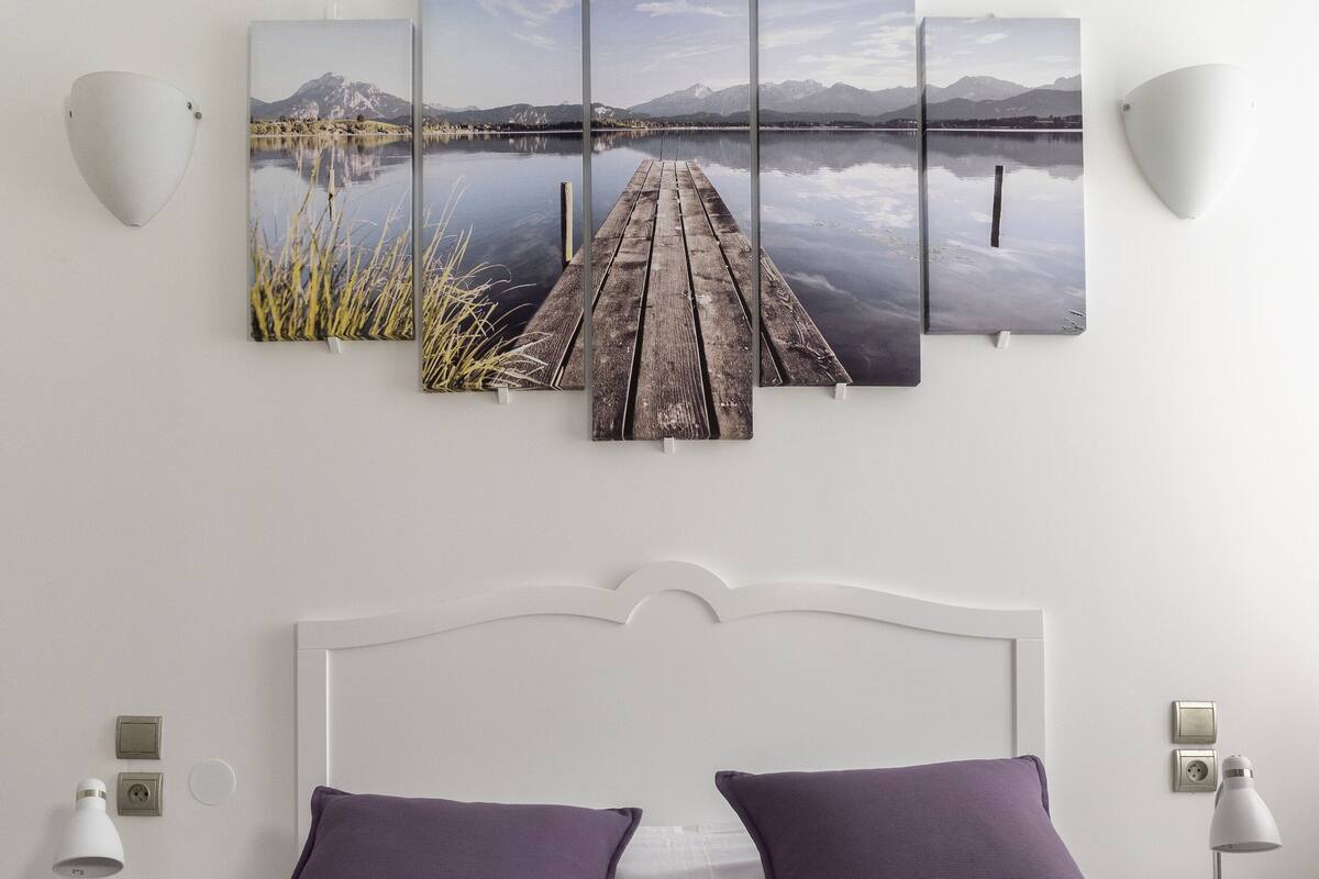 Un dormitorio con una cama, dos mesitas de noche, una lámpara de pared y una obra de arte de varios paneles sobre la cama.