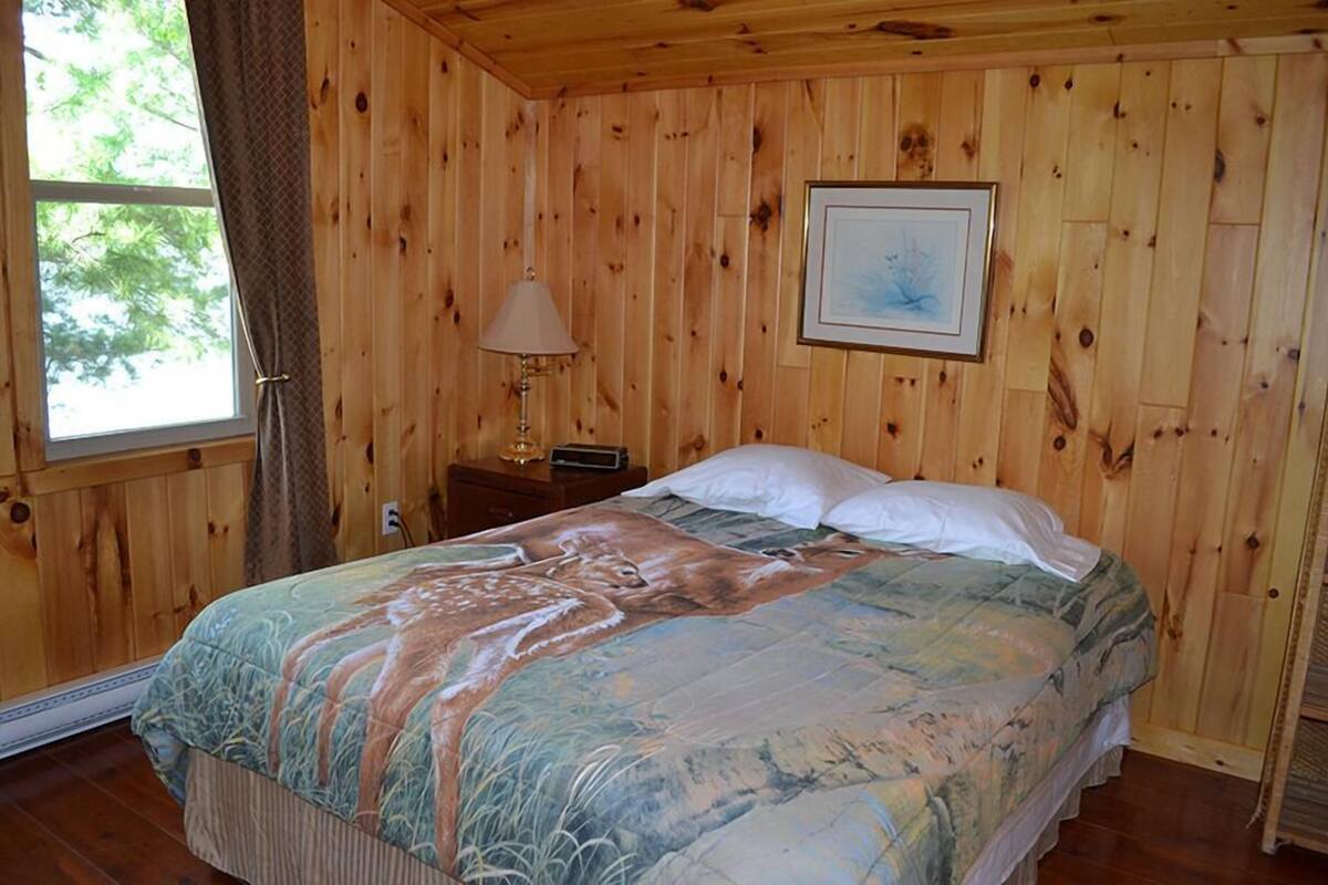 A wooden-paneled room with a bed, a nightstand, a lamp, and a framed picture on the wall.
