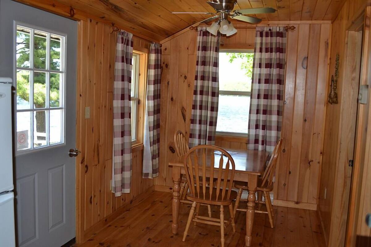 A wooden cabin interior with a dining table and chairs, plaid curtains, a ceiling fan, and a door with a window.