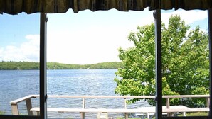 Une vue depuis une fenêtre donnant sur une terrasse en bois et des tables de pique-nique, avec un lac en contrebas, des arbres et des collines en arrière-plan.