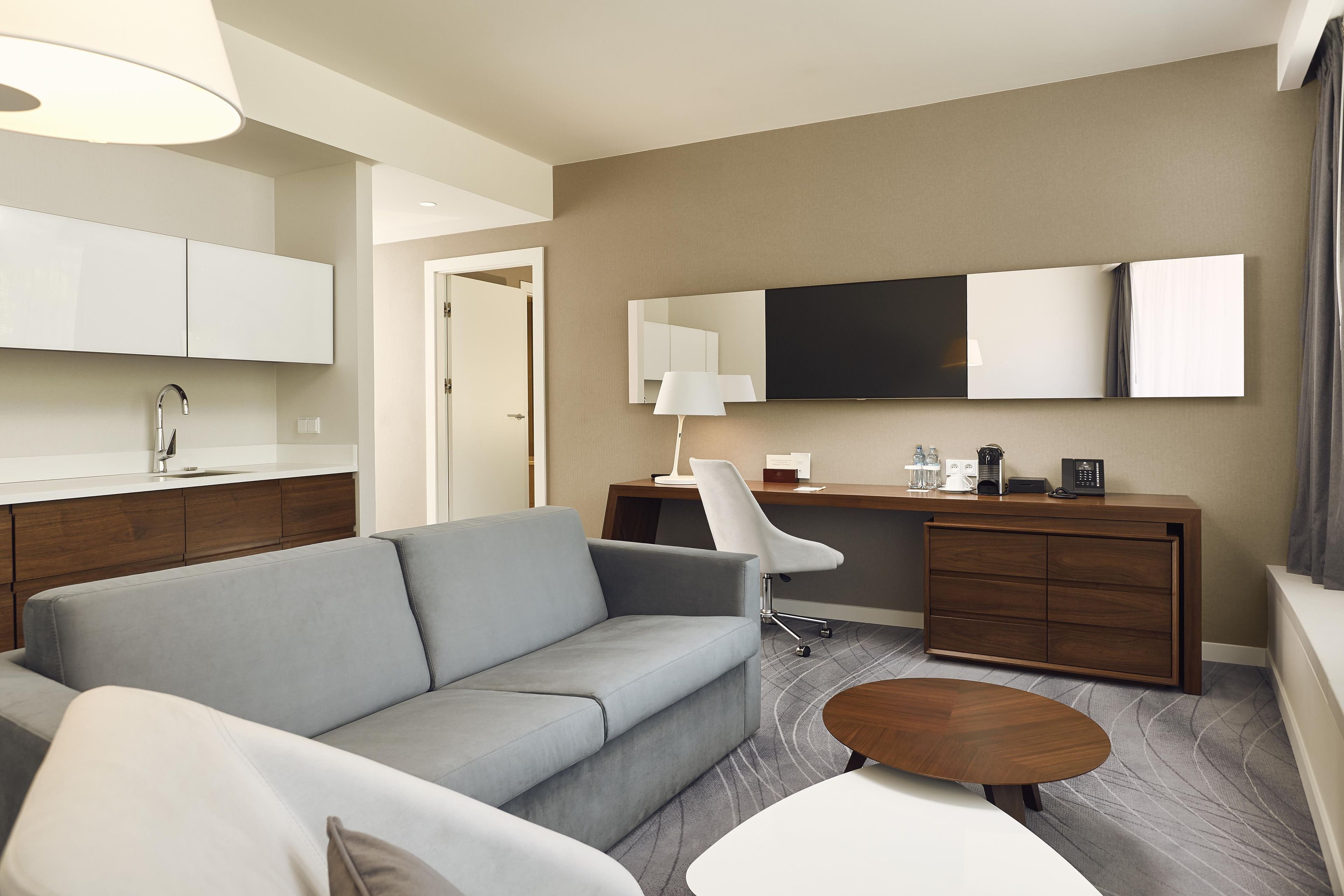 A hotel room with a sofa, a desk with a chair, a coffee machine, and a flat-screen TV.