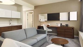 A hotel room with a sofa, a desk with a chair, a coffee machine, and a flat-screen TV.