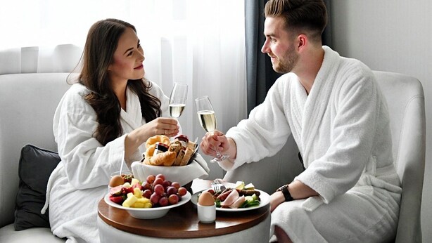 Et par i hvide badekåber sidder ved et rundt bord med morgenmad og glas vin.