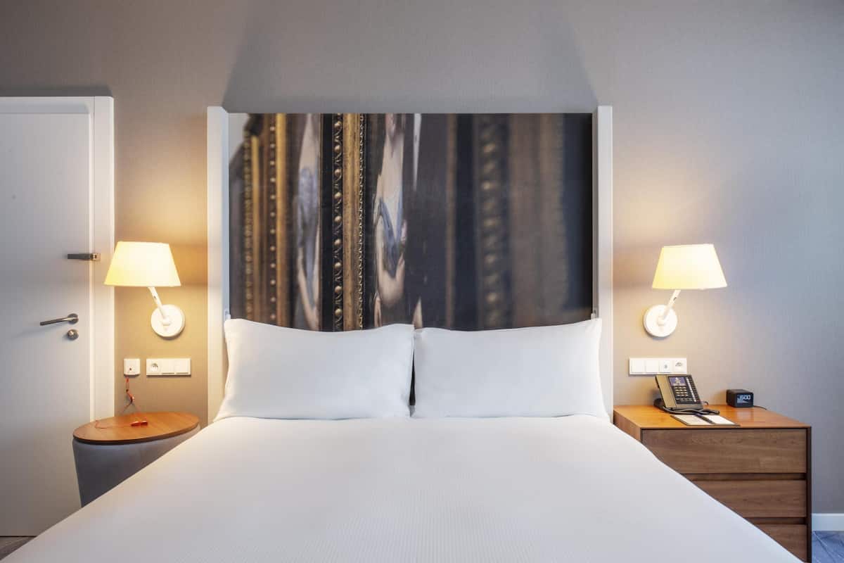 Habitación de hotel con cama, dos mesitas de noche con lámparas, teléfono y un interruptor de luz en la pared.