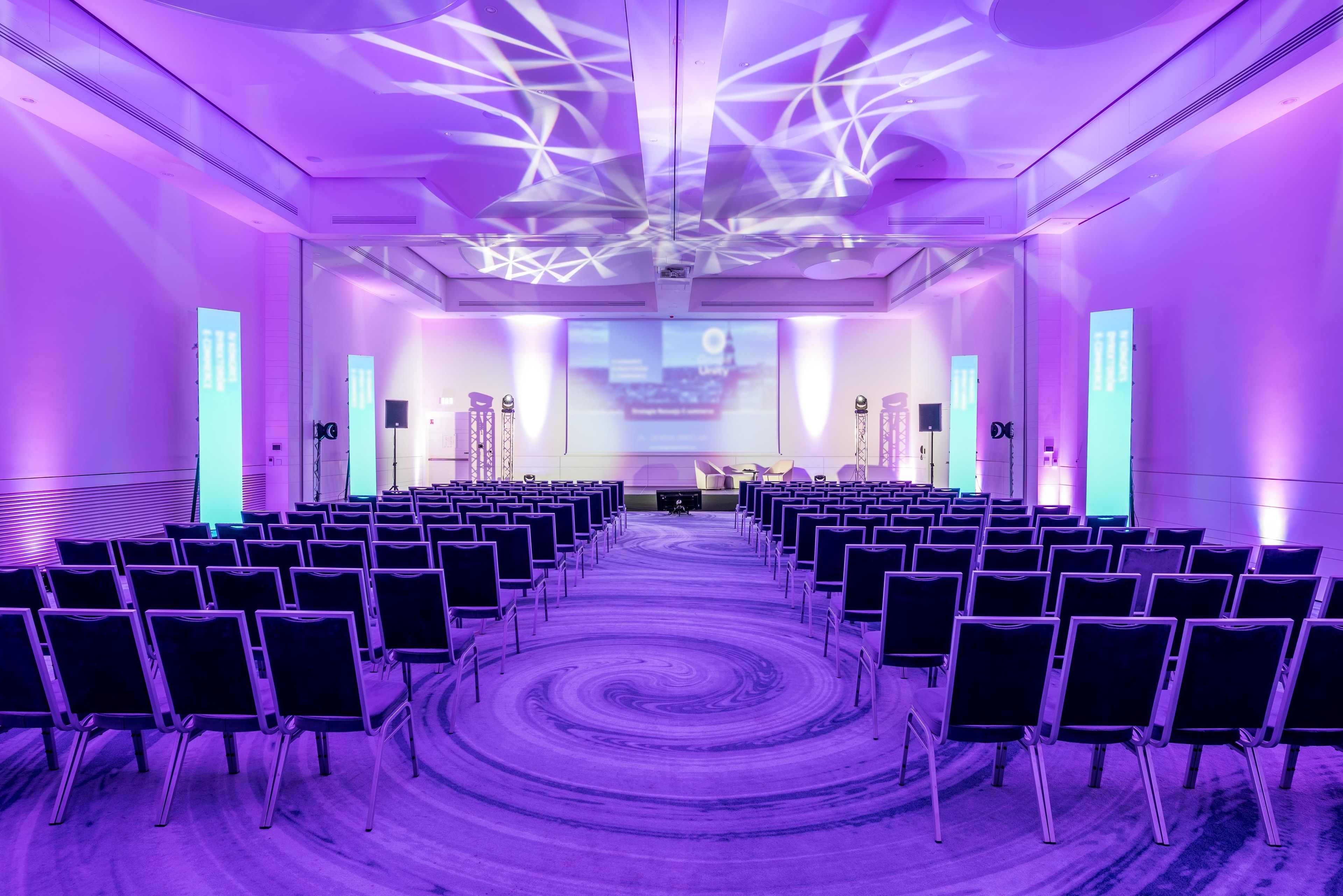 A conference room with rows of chairs facing a stage, equipped with a large screen and audio system.
