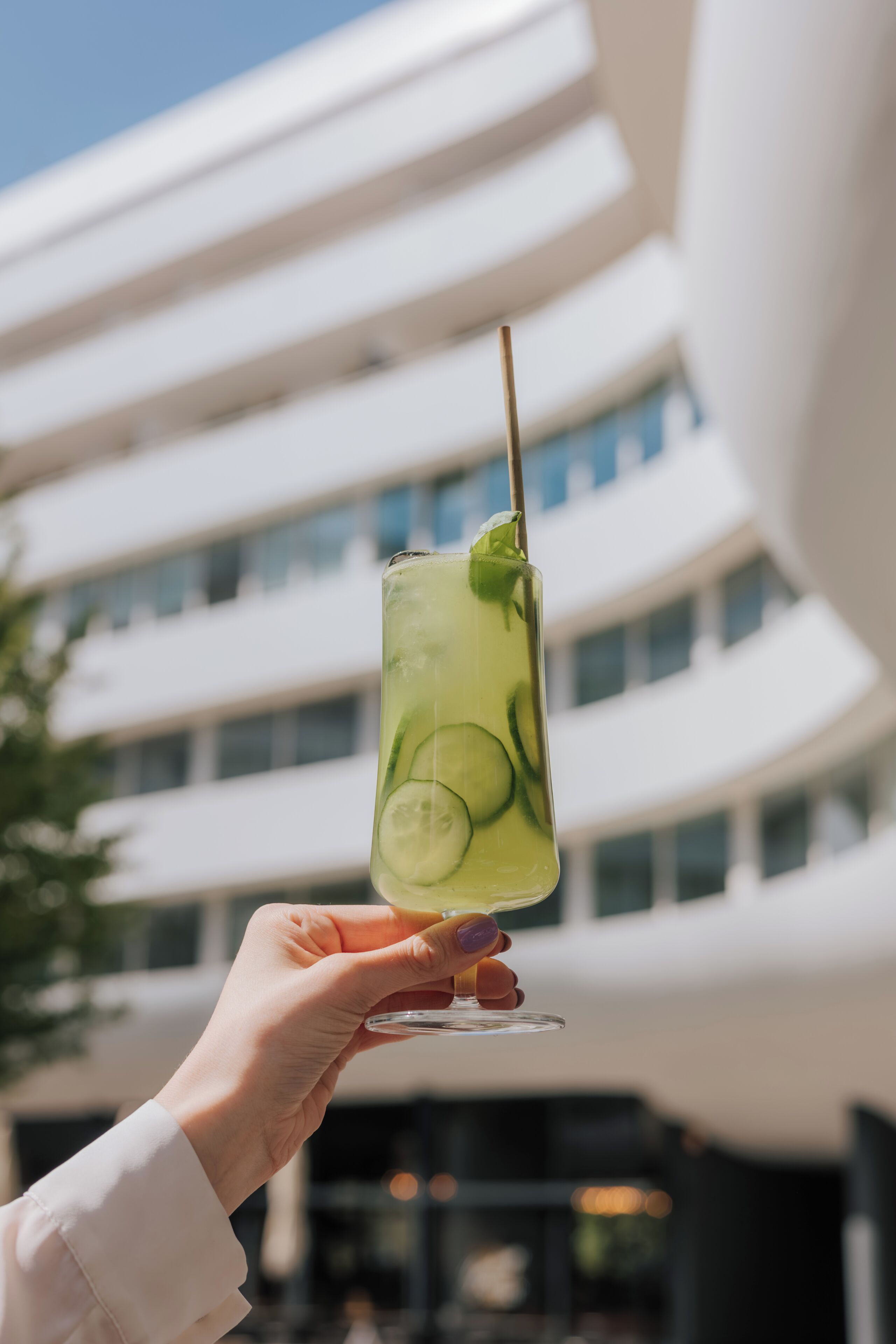 A hand holding a cocktail glass with a lime garnish, set against a modern building with multiple windows.