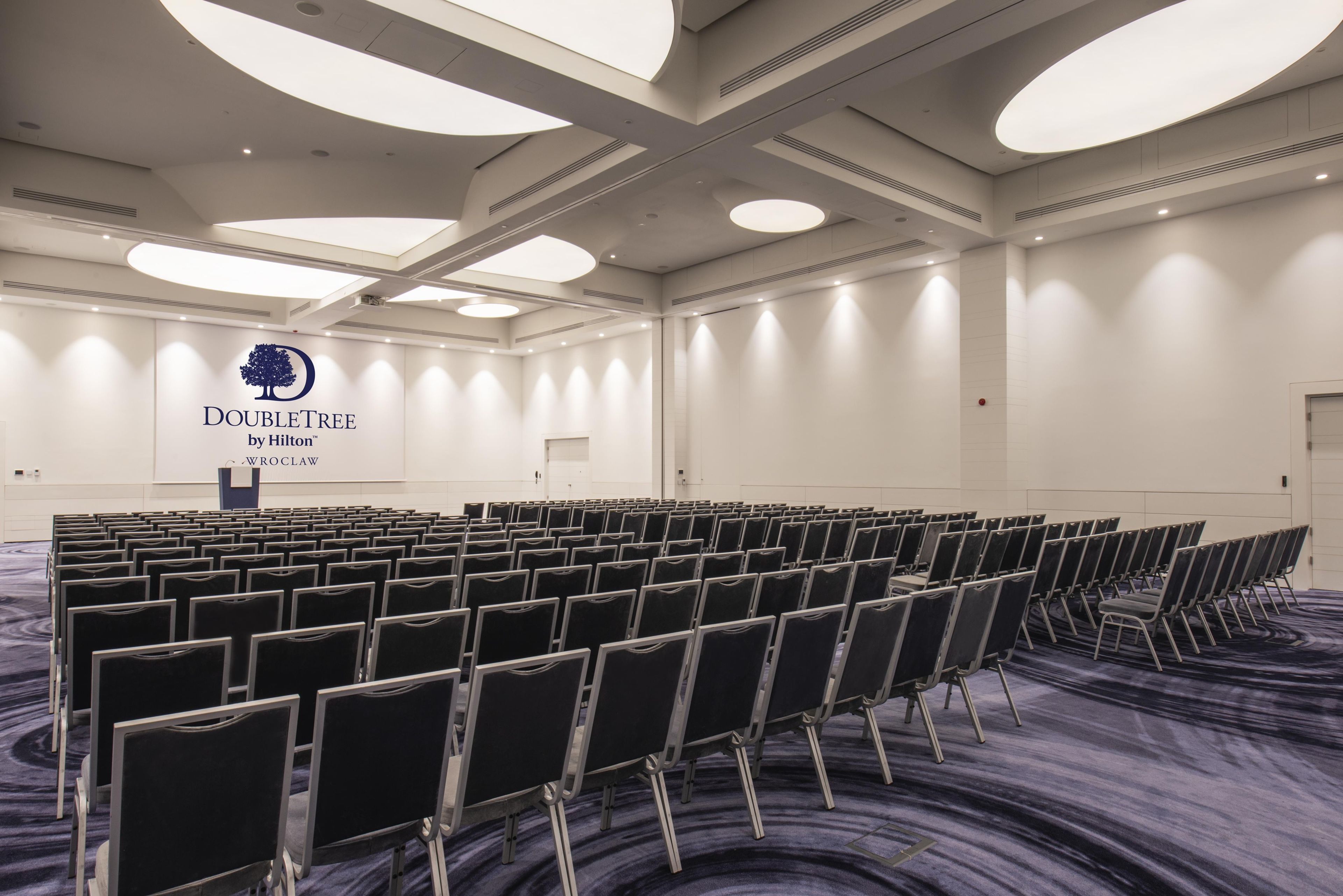 Uma sala de conferências com fileiras de cadeiras, um pódio com o logotipo da DoubleTree by Hilton e piso coberto por carpete.