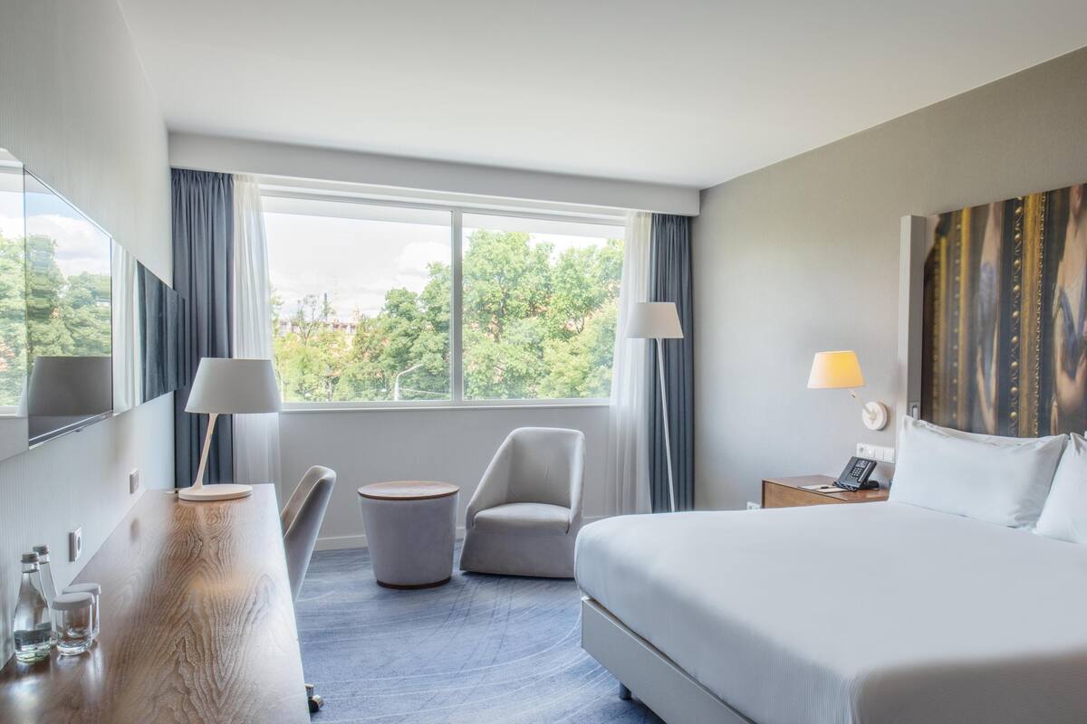 Habitación de hotel con una cama grande, un escritorio, una silla, una mesita, dos lámparas y vistas a árboles desde la ventana.