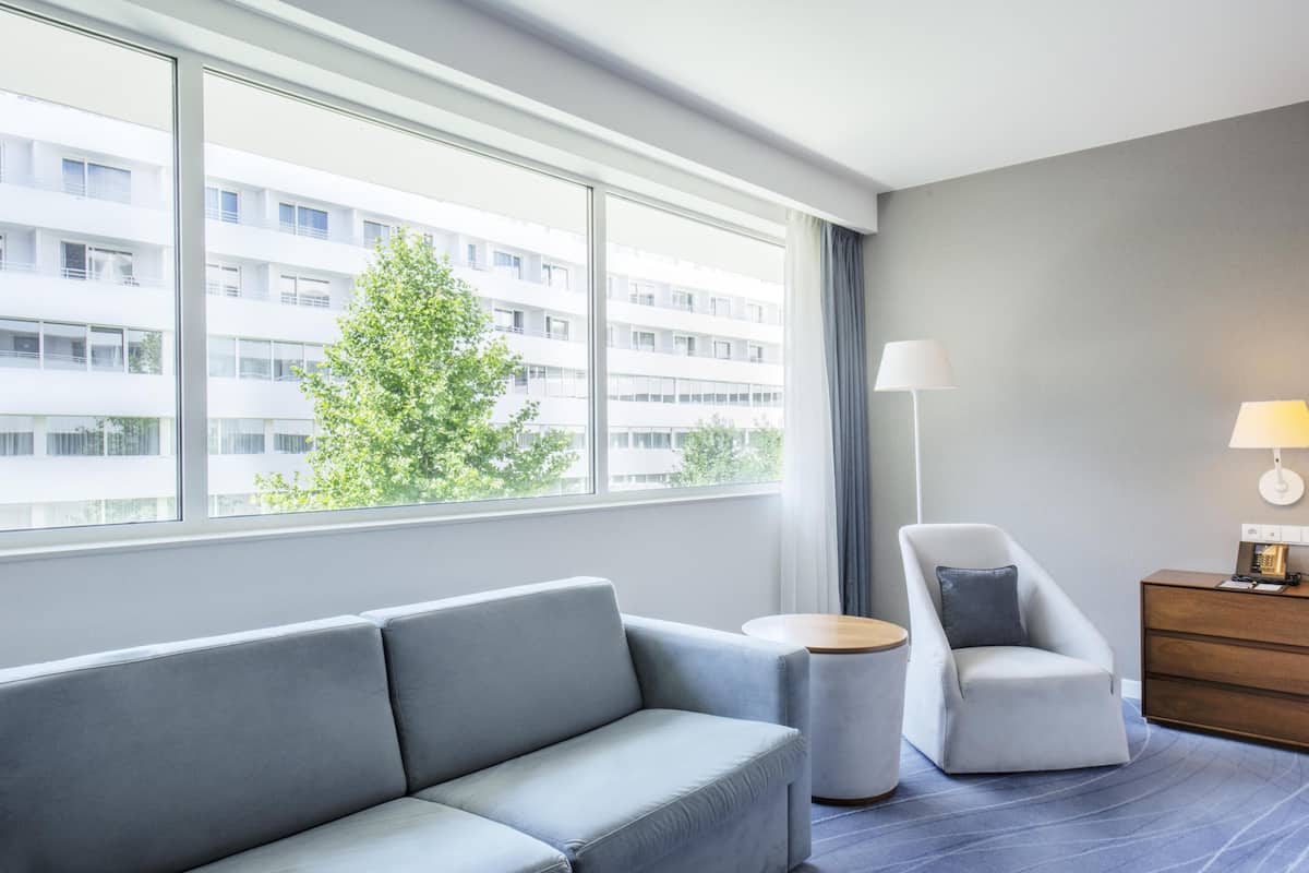 Una habitación de hotel moderna con un sofá gris, una mesita redonda, una silla, una lámpara y vistas a un edificio a través de amplios ventanales.