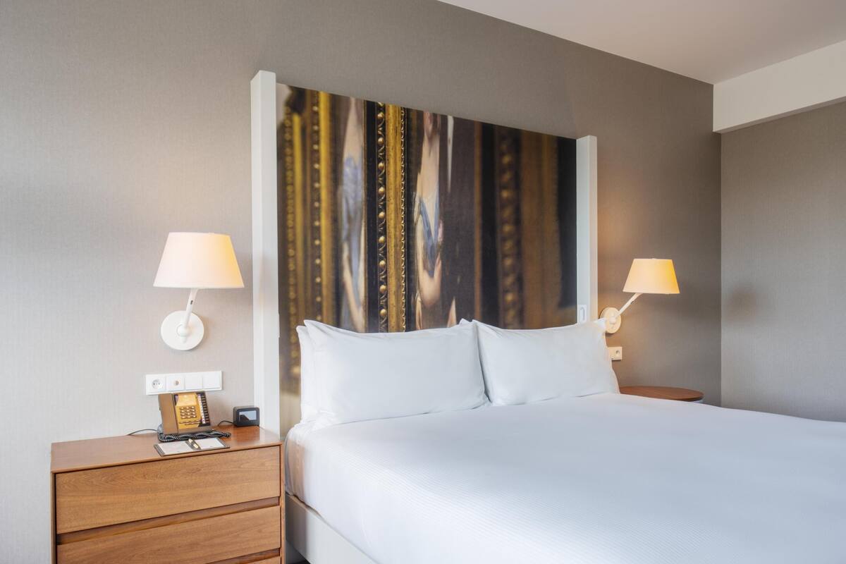 Habitación de hotel con cama, dos lámparas de noche, mesita con teléfono y una obra de arte en la pared.