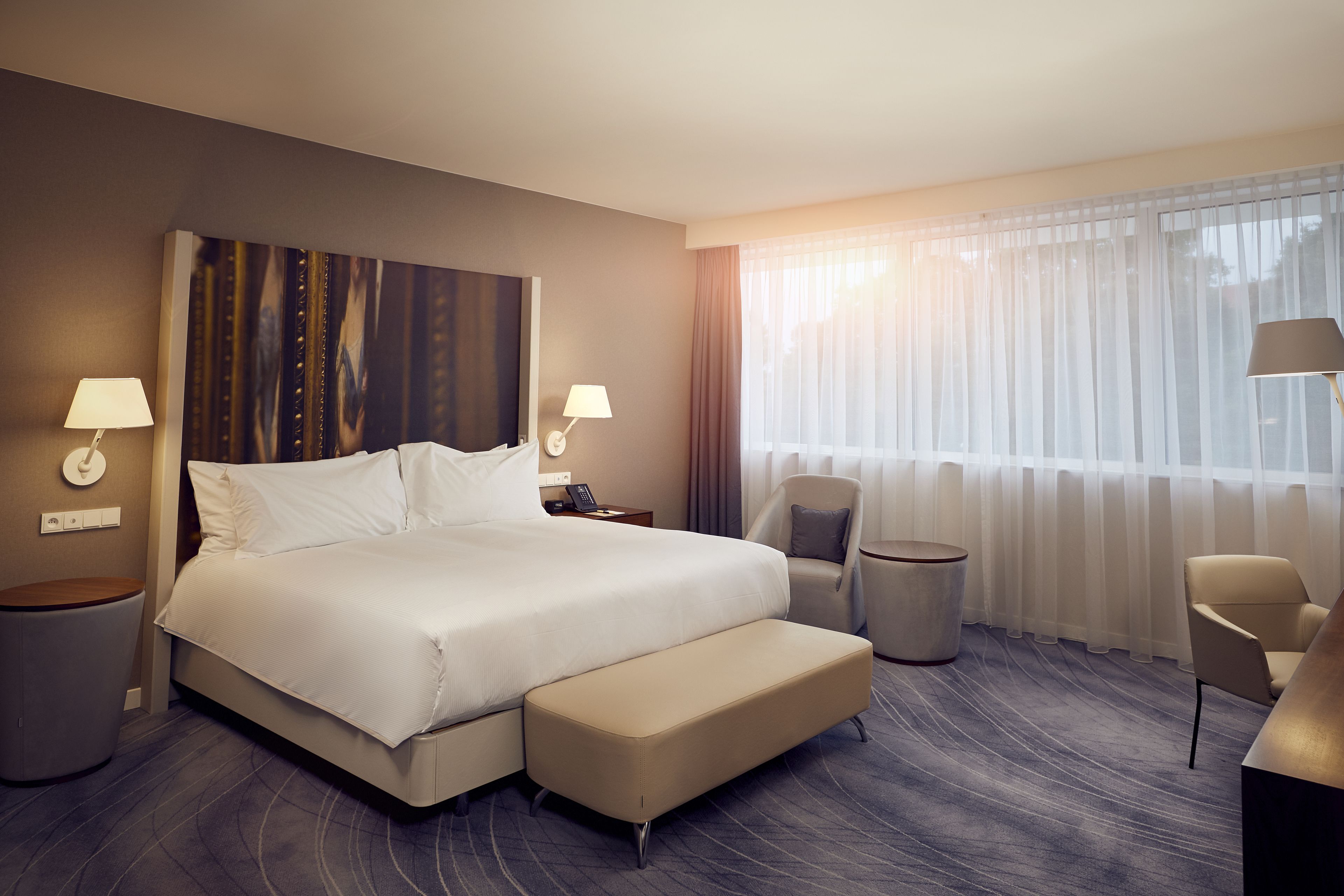 A modern hotel room with a large bed, a sitting area with a chair and a small table, a bedside lamp, and a view of the outdoors through sheer curtains.