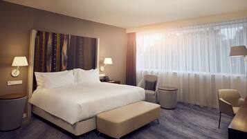 A modern hotel room with a large bed, a sitting area with a chair and a small table, a bedside lamp, and a view of the outdoors through sheer curtains.