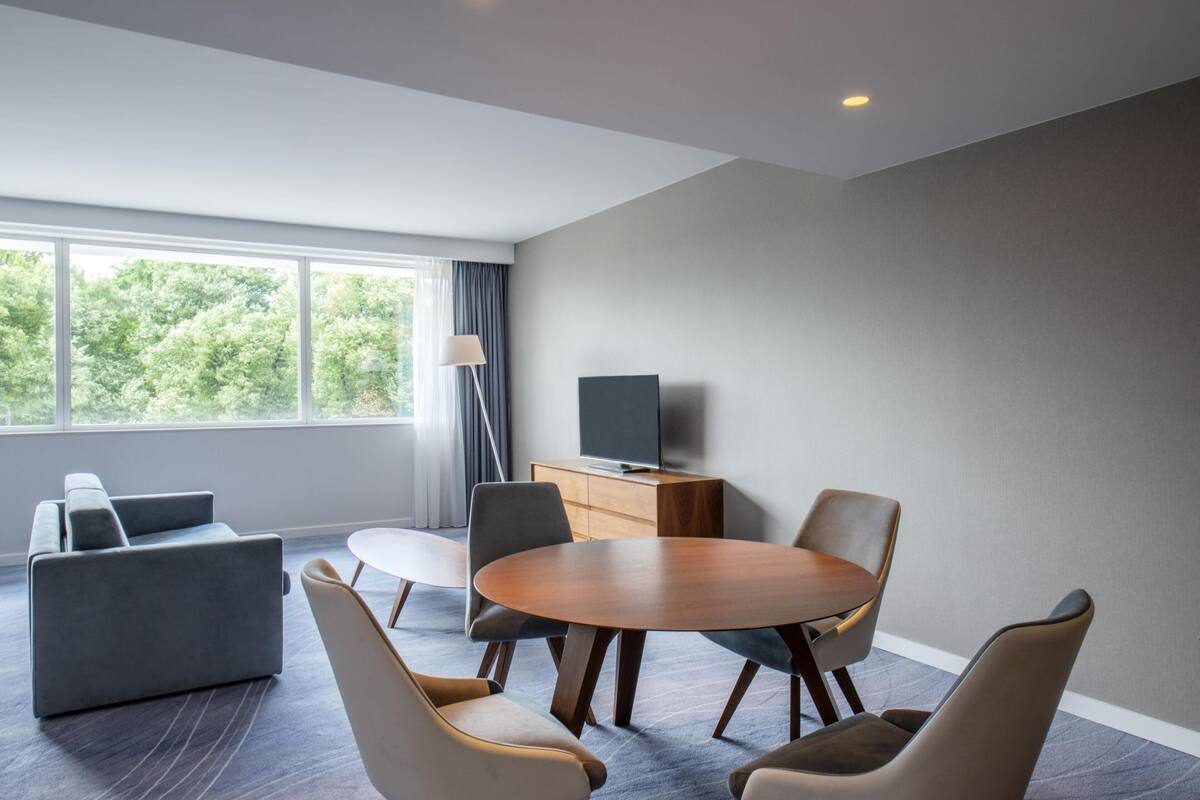 Una habitación moderna con una mesa redonda, sillas, un sofá, un televisor sobre un mueble de madera y una ventana con cortinas.