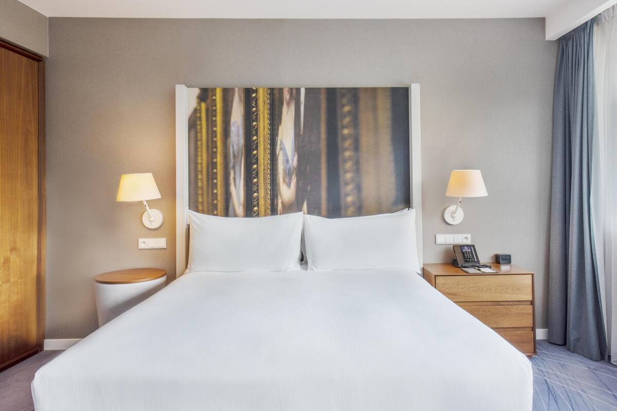 Una habitación de hotel con una cama grande, dos mesitas de noche con lámparas, un aparador de madera y un teléfono en la pared.