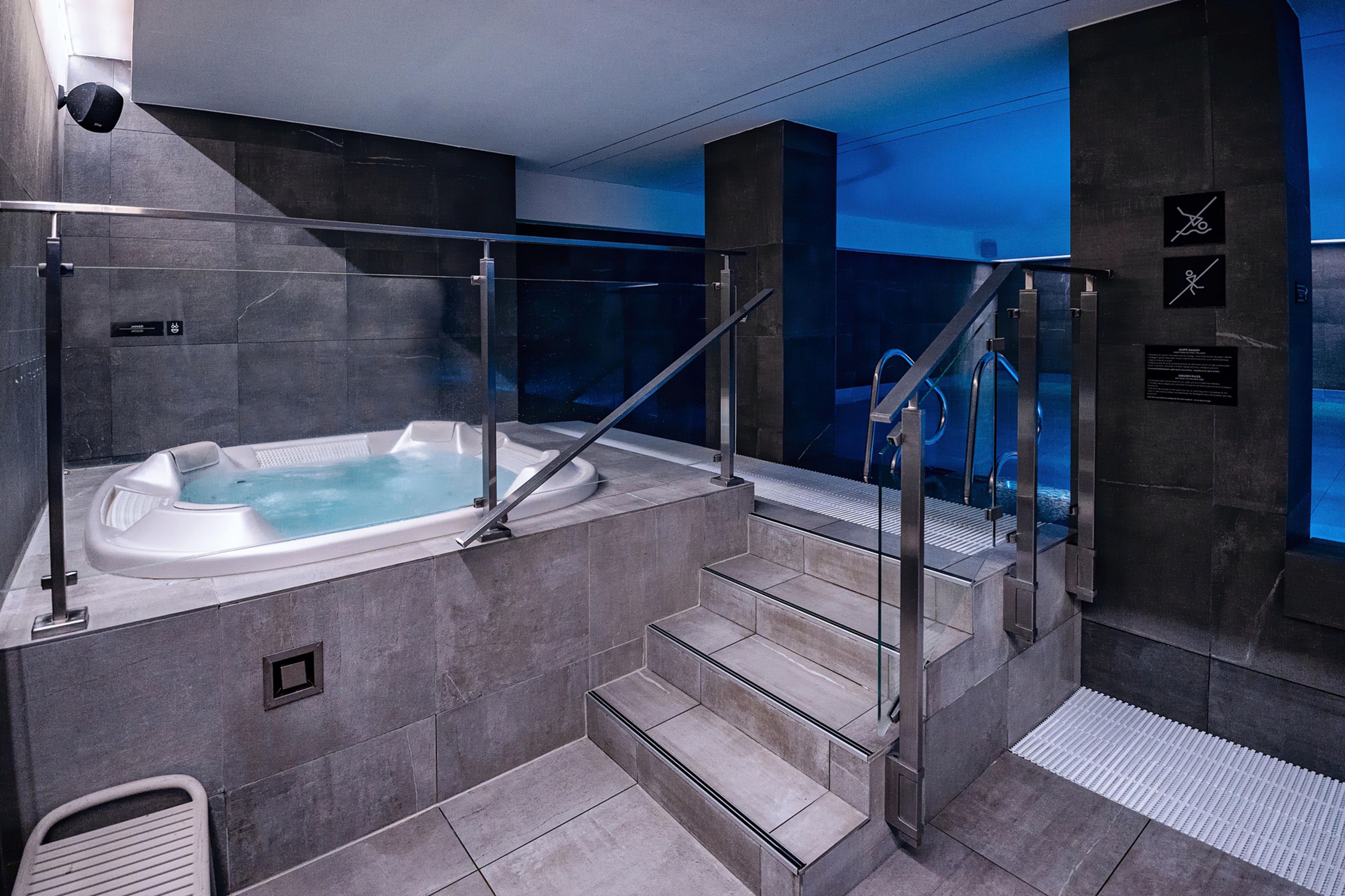 A modern indoor pool area with a hot tub, glass railings, and a staircase.