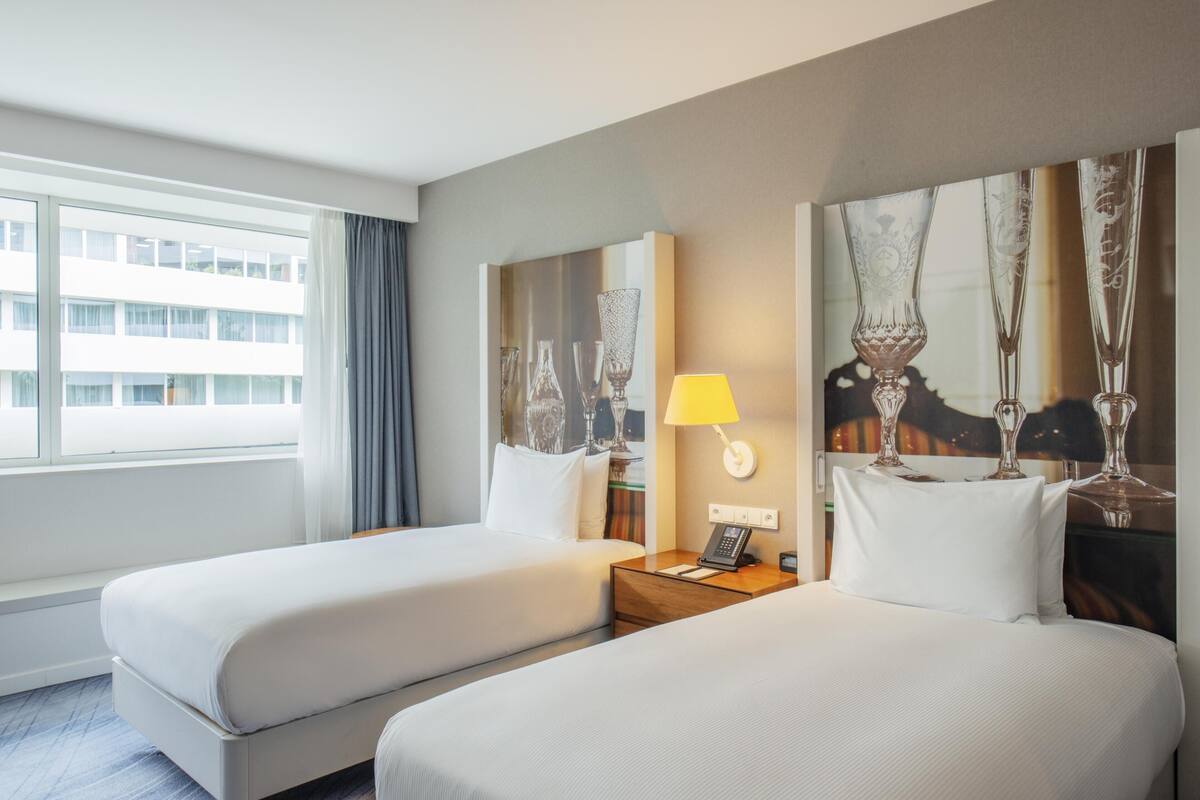 Habitación de hotel con dos camas, una ventana con cortinas, una lámpara y un cuadro de jarrones en la pared.