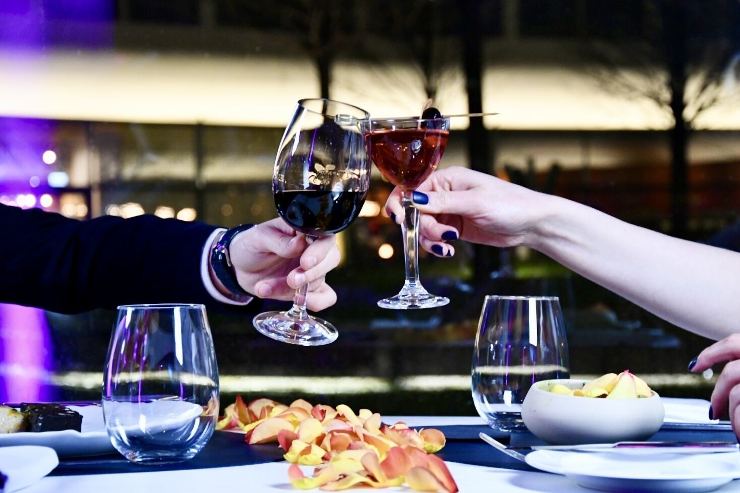 A dining table with two wine glasses, a bowl of fruit, and a plate of food.