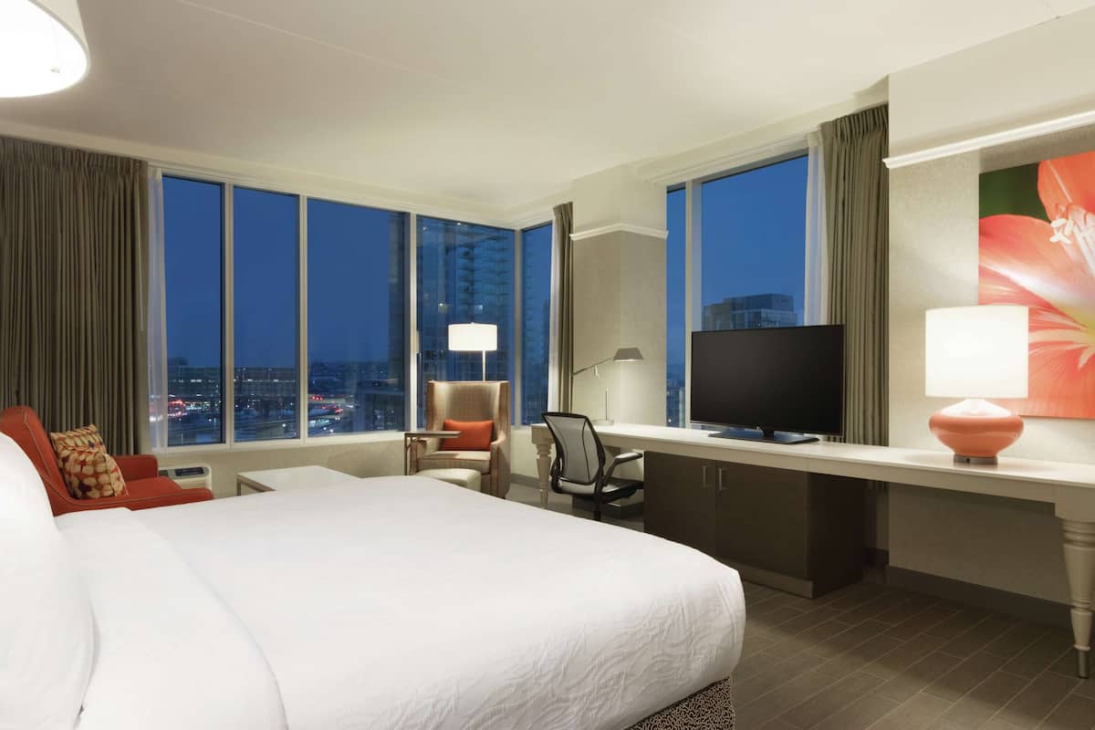 Une chambre d’hôtel avec un grand lit, un bureau avec une télévision, une chaise et une vue sur la ville la nuit.