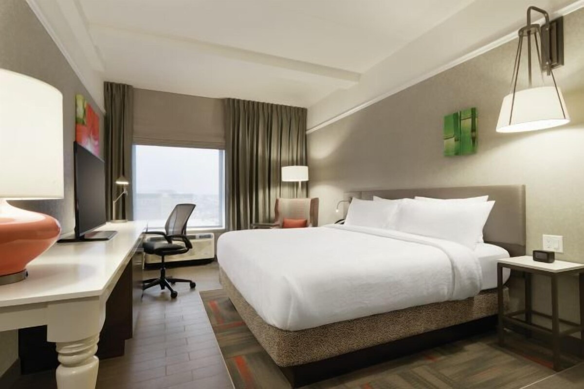 Habitación de hotel con una cama grande, un escritorio con silla, una ventana con cortinas y una lámpara.