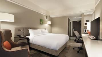 Una habitación de hotel con una cama grande, un escritorio con computadora, una silla, una mesita de noche y una lámpara.