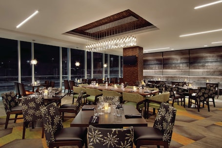 A modern dining area with multiple tables and chairs, large windows, and a central chandelier.. Homewood Suites by Hilton Calgary Downtown