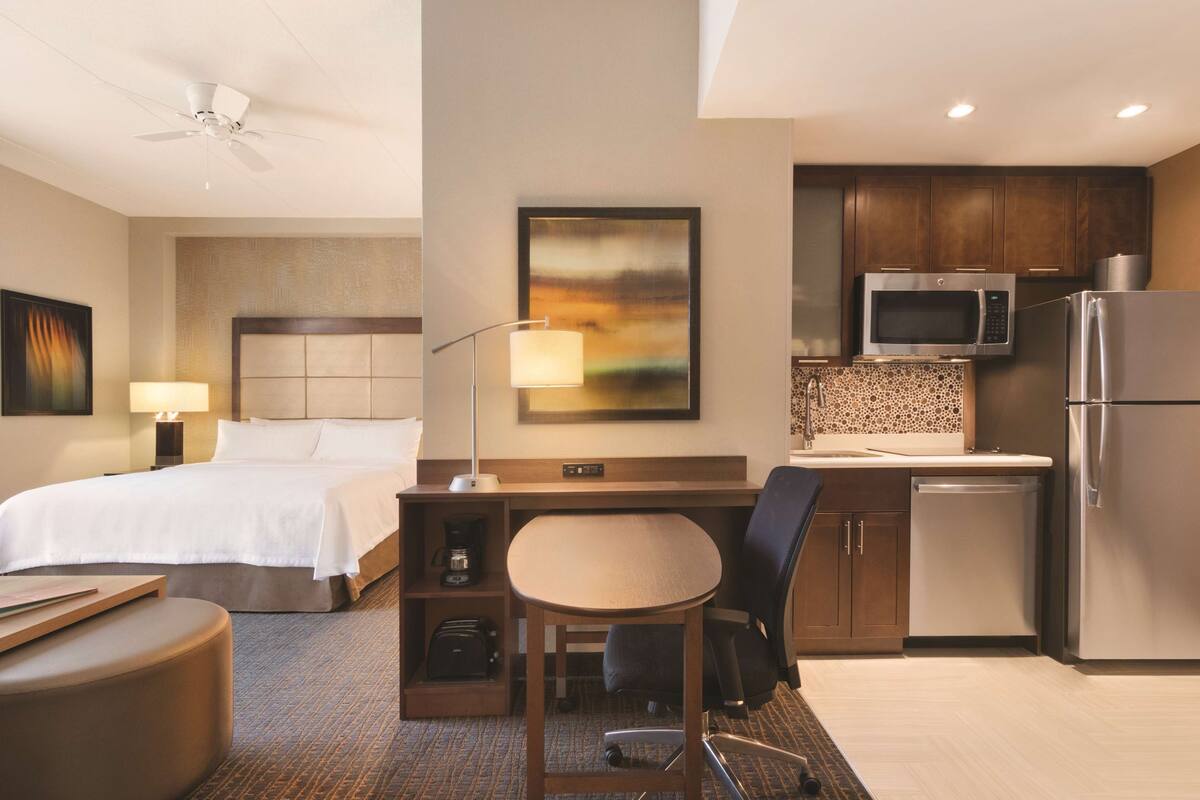 Una habitación de hotel con cama, escritorio, silla, kitchenette y ventilador de techo.