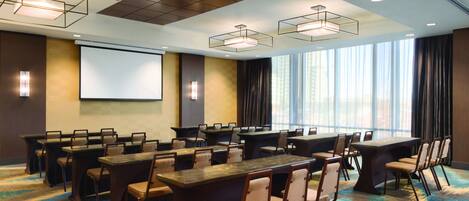A conference room with multiple tables and chairs, a projector screen, and large windows with curtains.