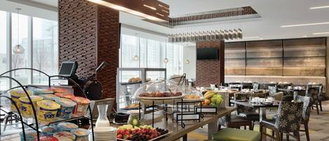 A modern hotel dining area with a buffet setup, seating arrangements, and a large TV screen.
