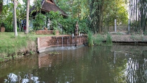 A serene pond with a small bridge, surrounded by lush greenery and a traditional building with a tiled roof. - Taladya Chiang Mai Homestay (Doi Saket)