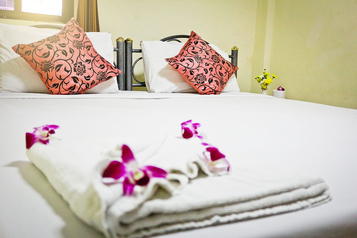 Una cama bien hecha con dos almohadas decorativas, una toalla doblada con flores moradas y un pequeño florero con flores amarillas sobre una mesita de noche.