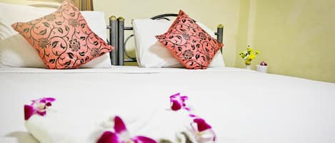 A neatly made bed with two decorative pillows, a folded towel with purple flowers, and a small vase with yellow flowers on a bedside table.