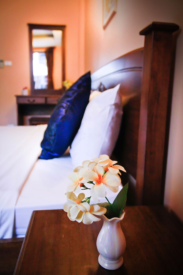 Una cama con ropa de cama blanca, una almohada azul y un florero con flores blancas sobre una mesita de noche de madera.