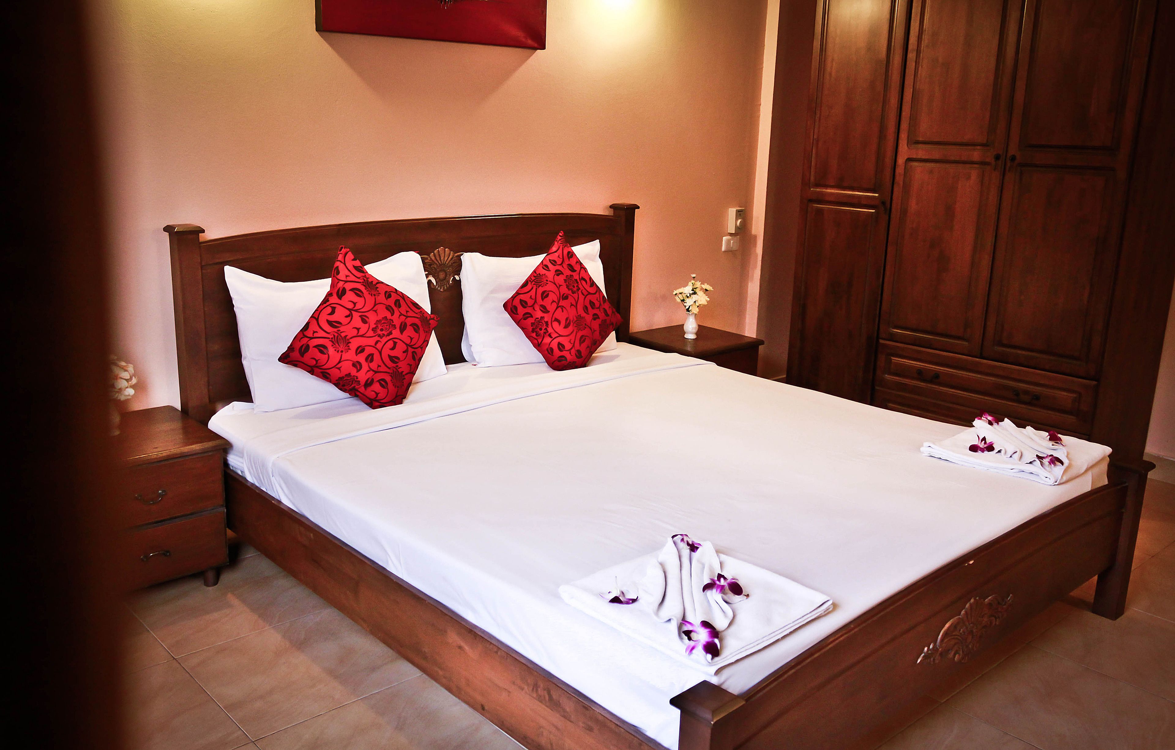 A neatly made bed with white linens and red patterned pillows, a wooden nightstand with a vase of flowers, and a large wooden wardrobe.