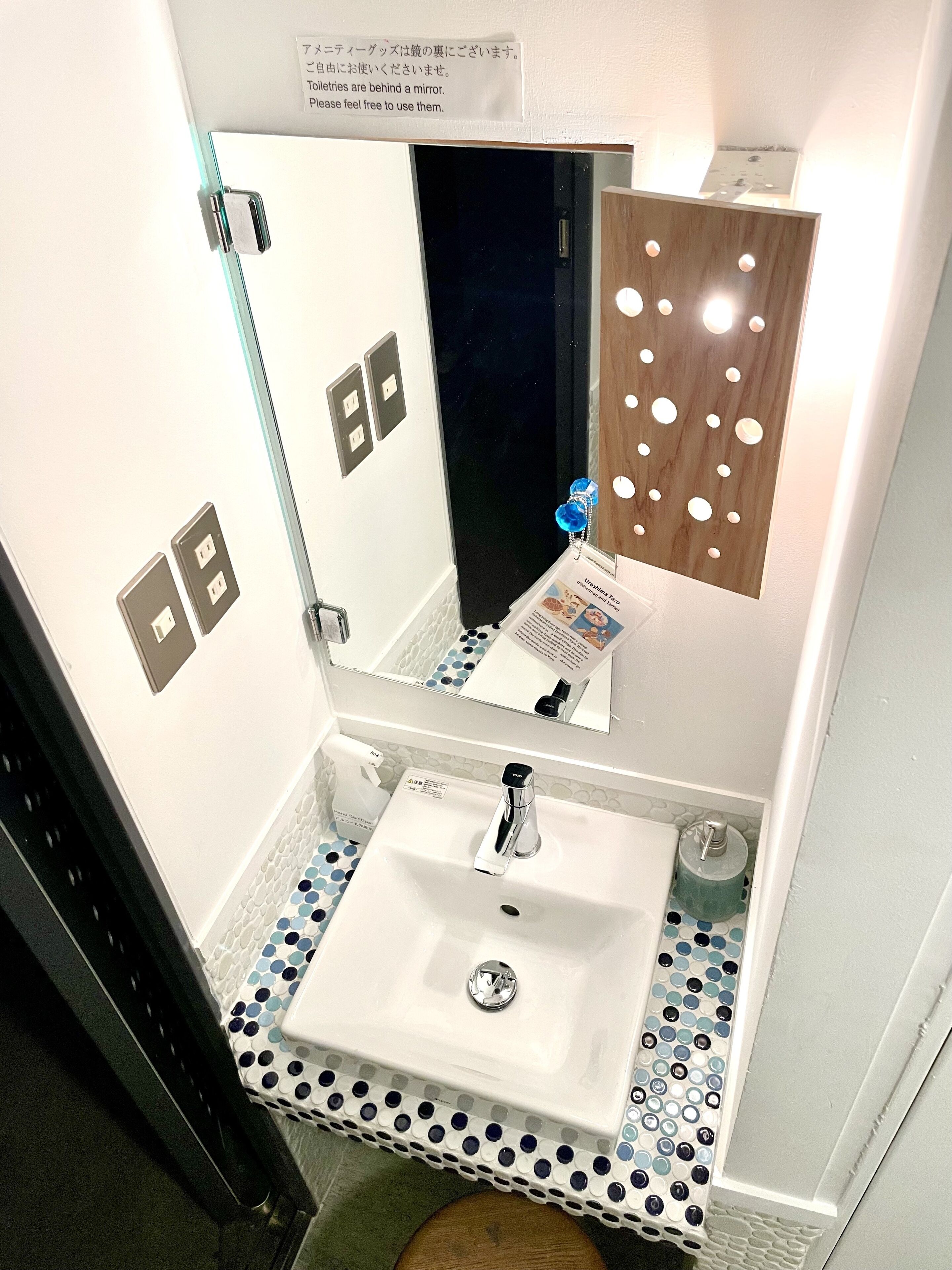 A bathroom with a white sink, a mirror, and a wooden panel with circular cutouts. There are two electrical outlets on the wall and a sign above the mirror.