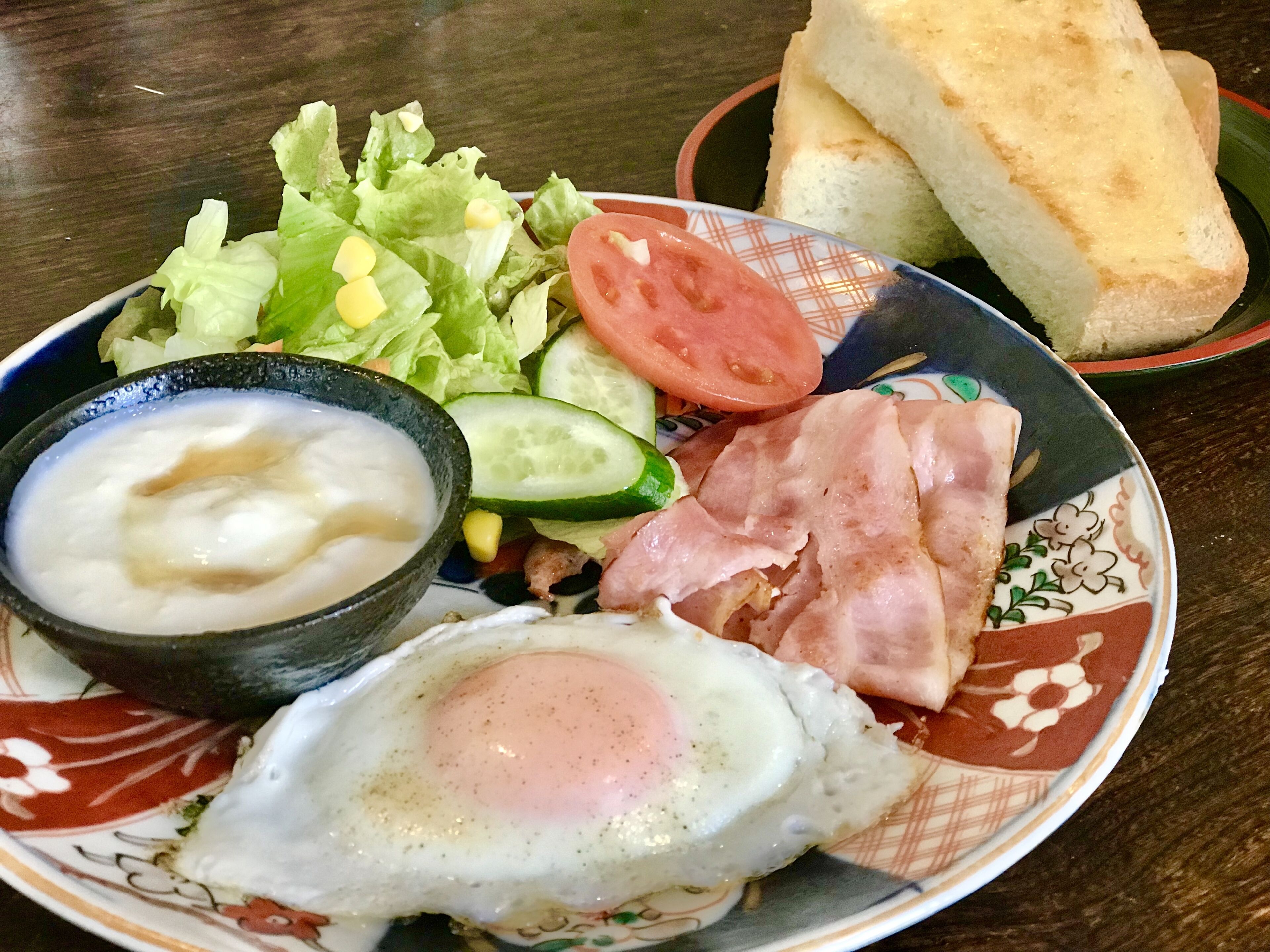 A plate with a fried egg, ham, lettuce, tomato, cucumber, and a bowl of soup.