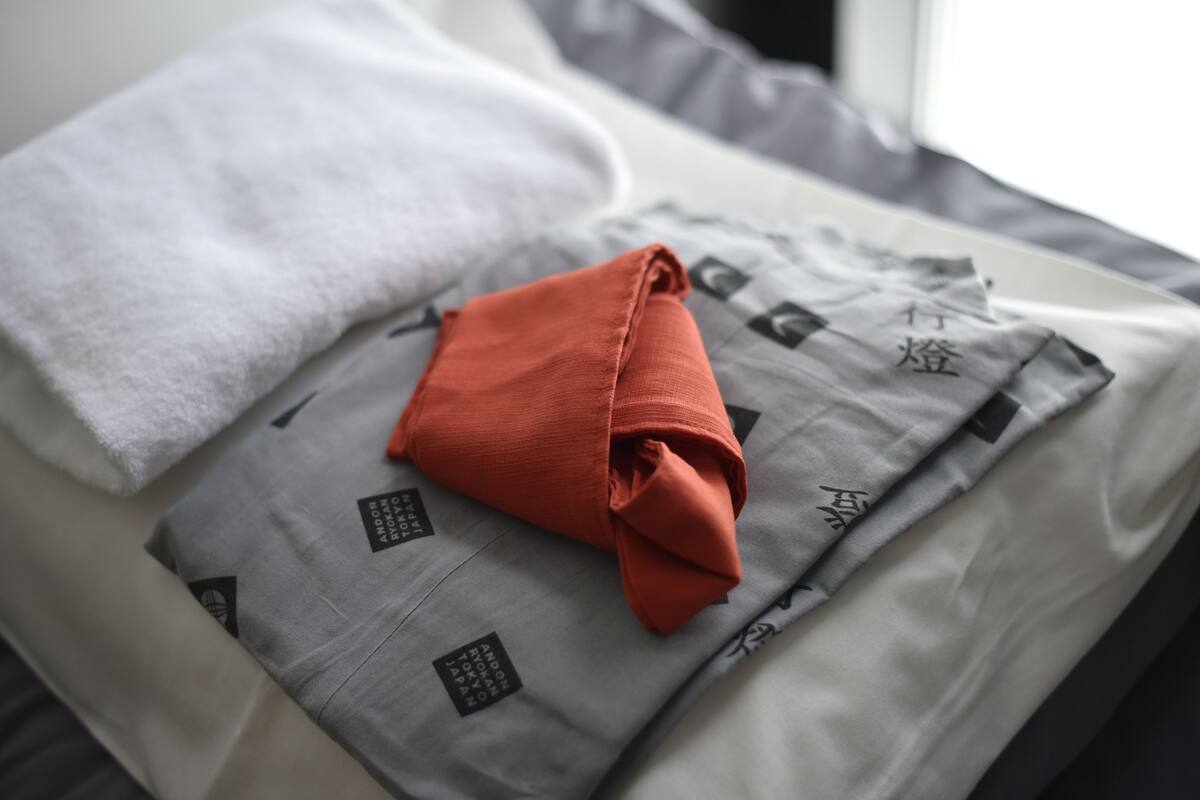 A neatly made bed with a grey and white patterned bedspread, a folded red towel, and a white pillow.