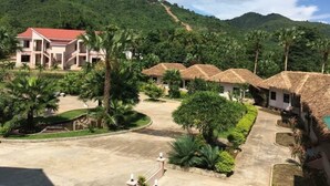 Un resort con un edificio grande y varias estructuras de techo de paja, rodeado de abundante vegetación y colinas.