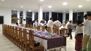 Una larga mesa de comedor con sillas, personas de pie conversando y una estructura de techo de paja al fondo.