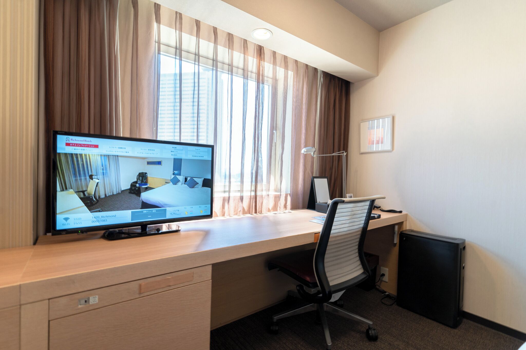 A hotel room with a desk, a computer, a chair, a lamp, and a window with curtains.