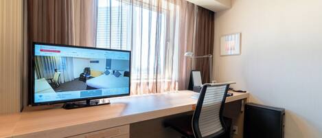 A hotel room with a desk, a computer, a chair, a lamp, and a window with curtains.