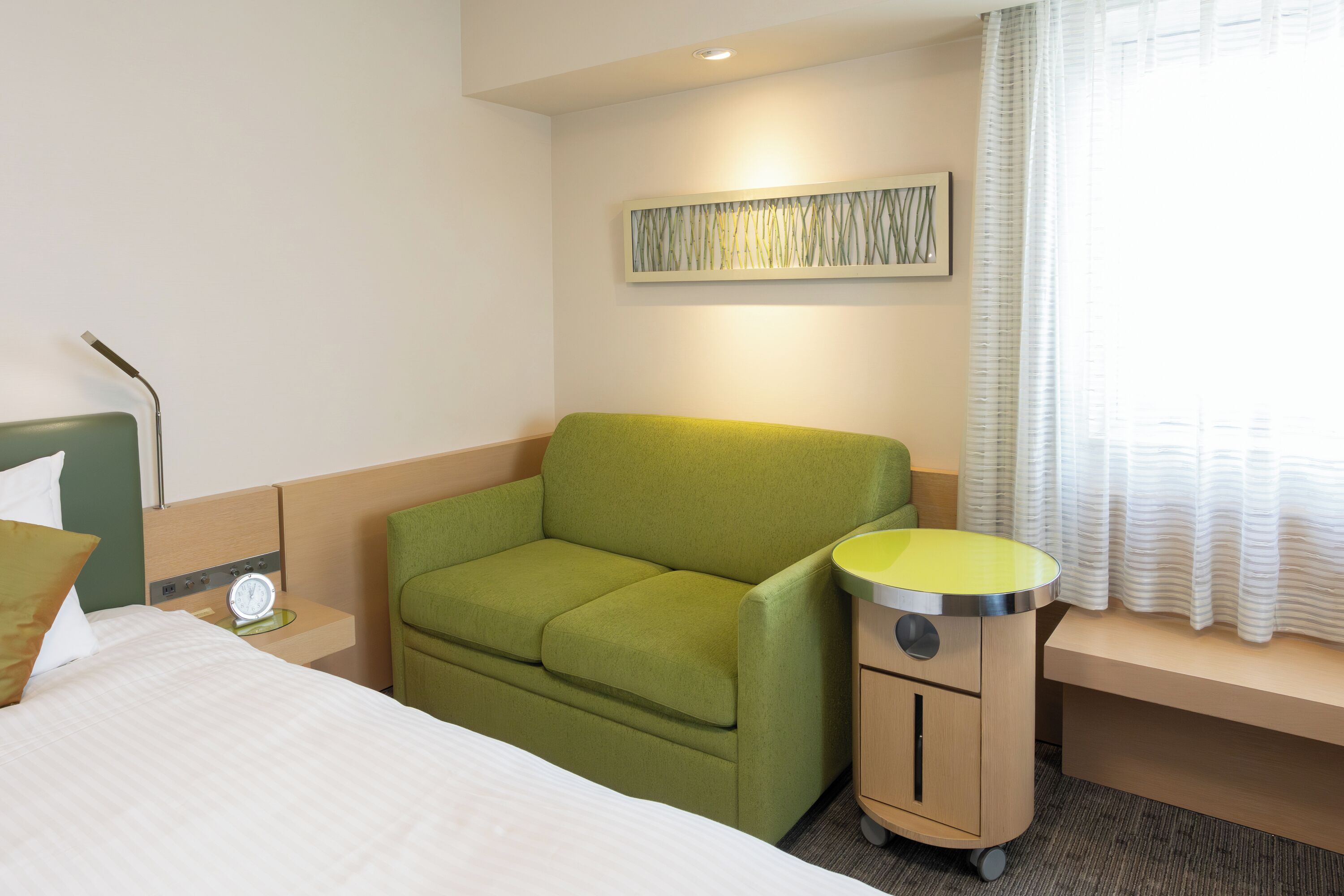 A hotel room with a bed, a green armchair, a small bedside table with a lamp, a wall-mounted artwork, and a window with curtains.
