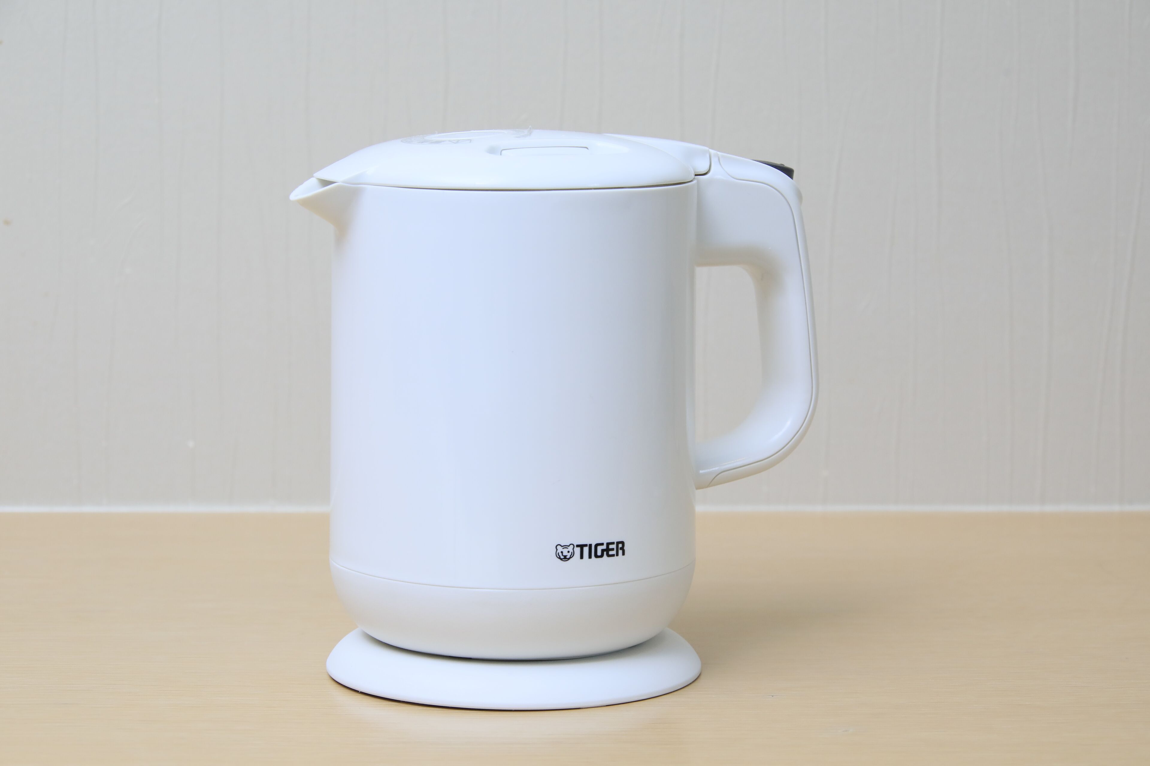 A white electric kettle with a handle and a lid, placed on a wooden surface.