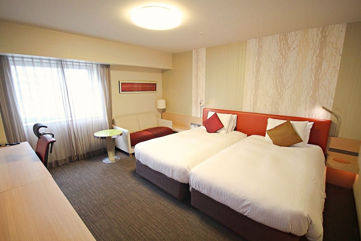 Een hotelkamer met twee bedden, een bureau, een stoel, een kleine tafel en een raam met gordijnen.