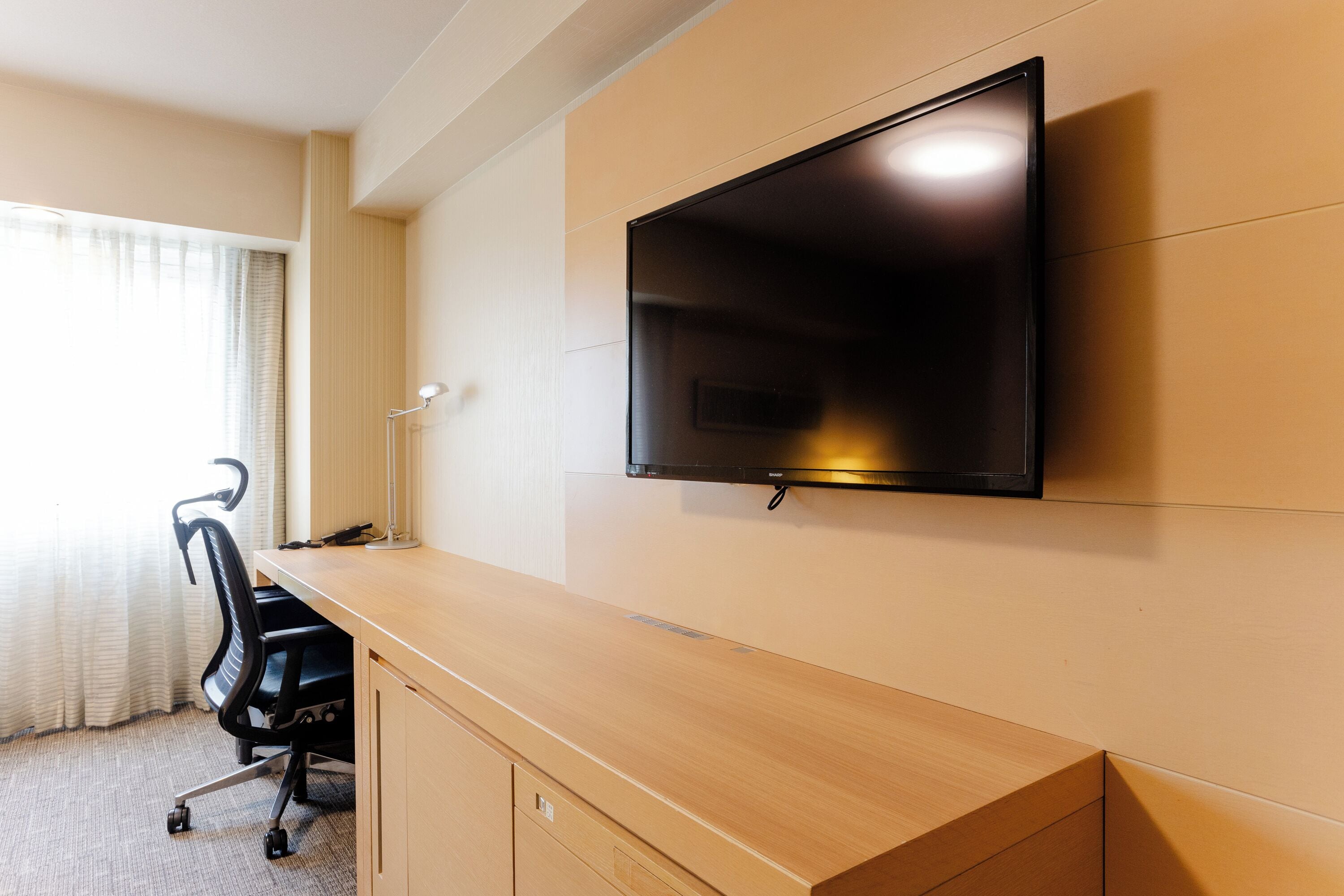 Une chambre d’hôtel avec un bureau en bois, un fauteuil de bureau noir, un téléviseur à écran plat fixé au mur et une fenêtre avec des rideaux.