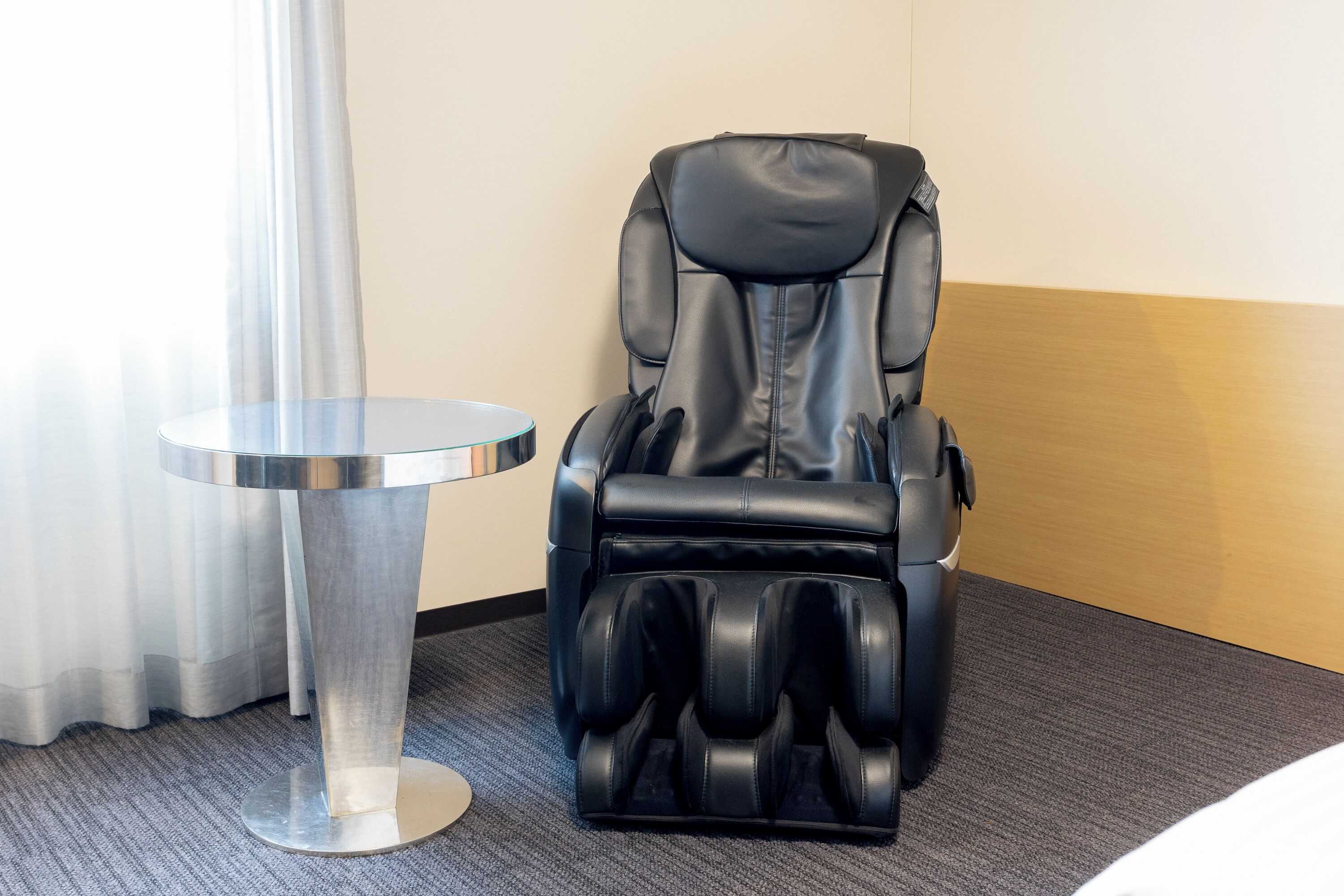 Uma cadeira de massagem num quarto de hotel, com uma pequena mesa redonda e uma cortina.