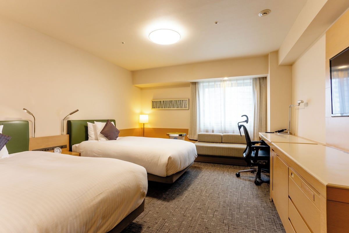 Een hotelkamer met twee bedden, een bureau, een stoel, een lamp en een raam met gordijnen.