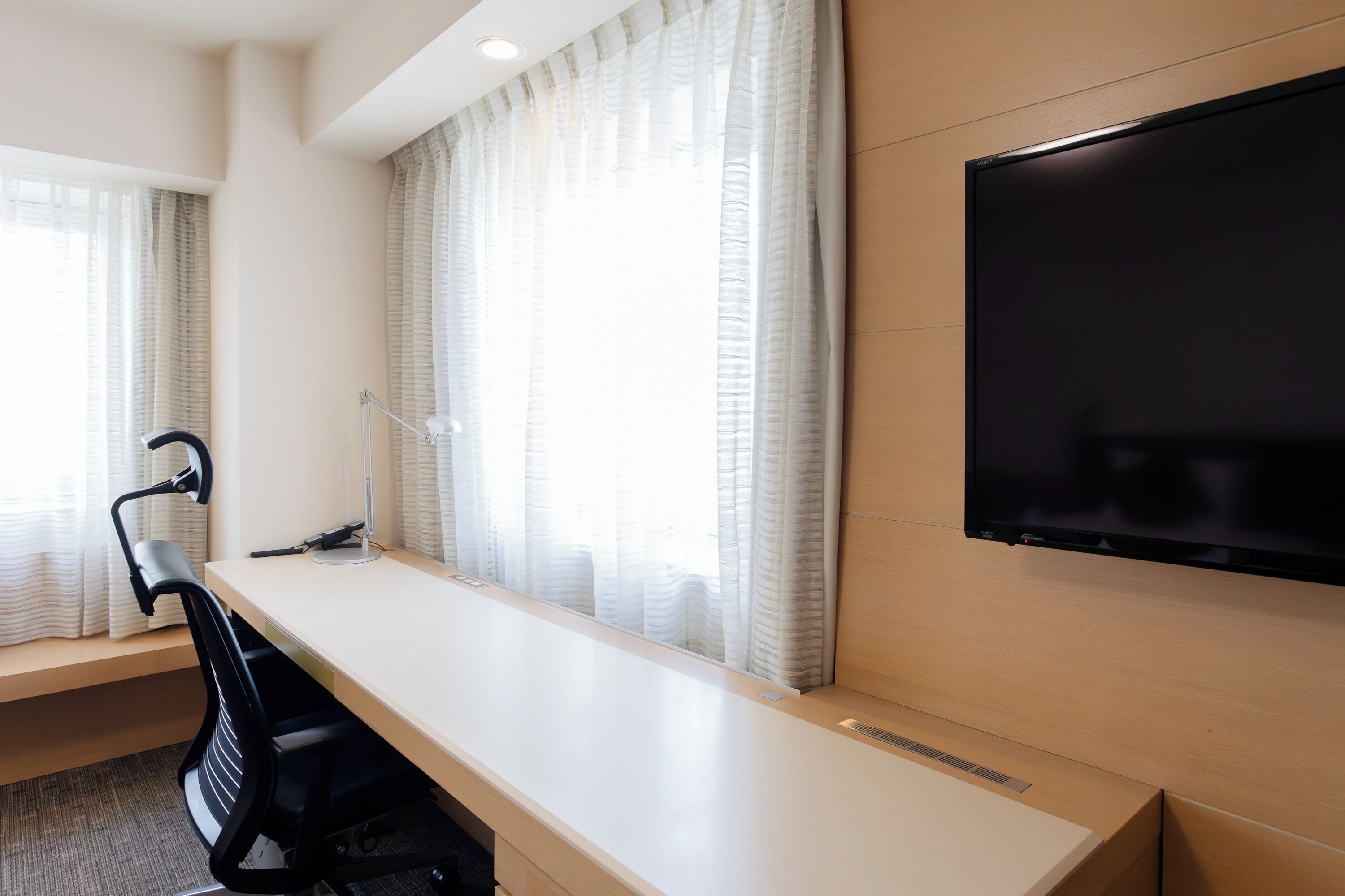 A modern hotel room with a large desk, a flat-screen TV mounted on the wall, and large windows with sheer curtains.